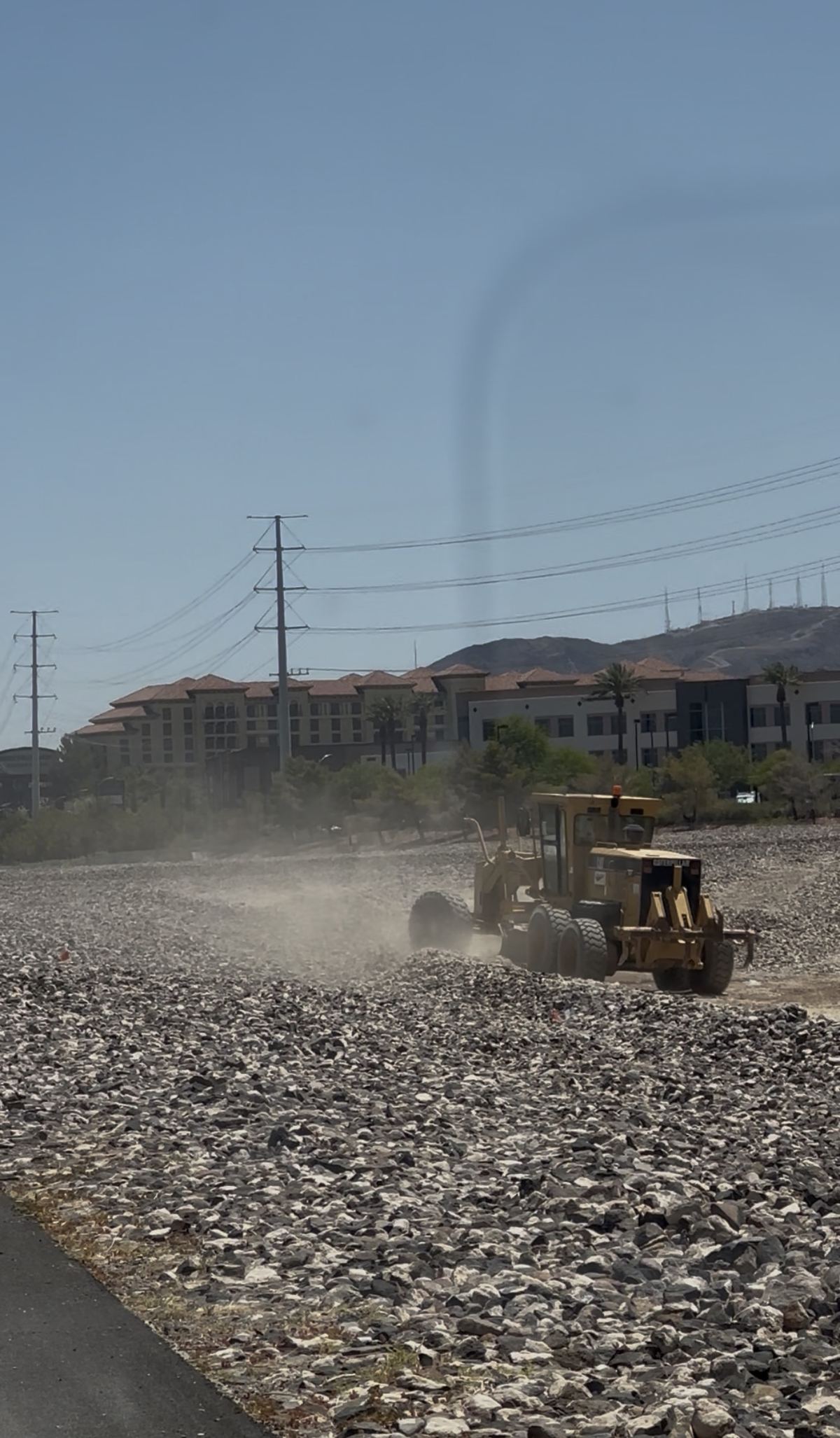 CC-215 grader with visible dust plume, hotel in background
