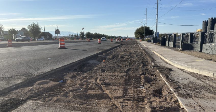 Torn-up median and disturbed soil beside an active traffic lane on Boulder Highway, with construction barrels along the roadway
